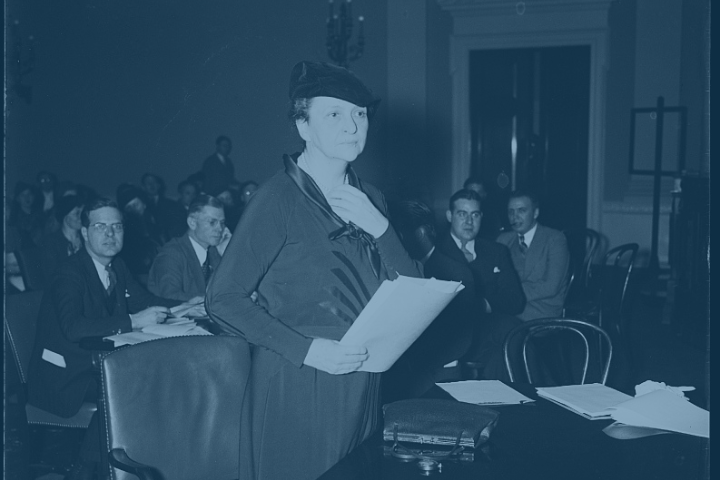 A woman holding a stack of papers stands in front of a desk. Behind her are rows of men and women in suits sitting at long tables.