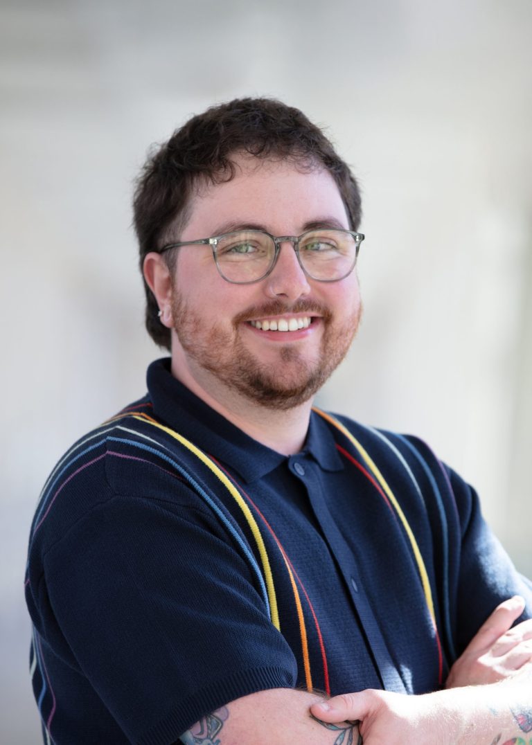 Professional headshot of VPLC employee Ezra Halstead, who has short curly brown hair and is wearing a navy button up shirt with rainbow stripes on it.