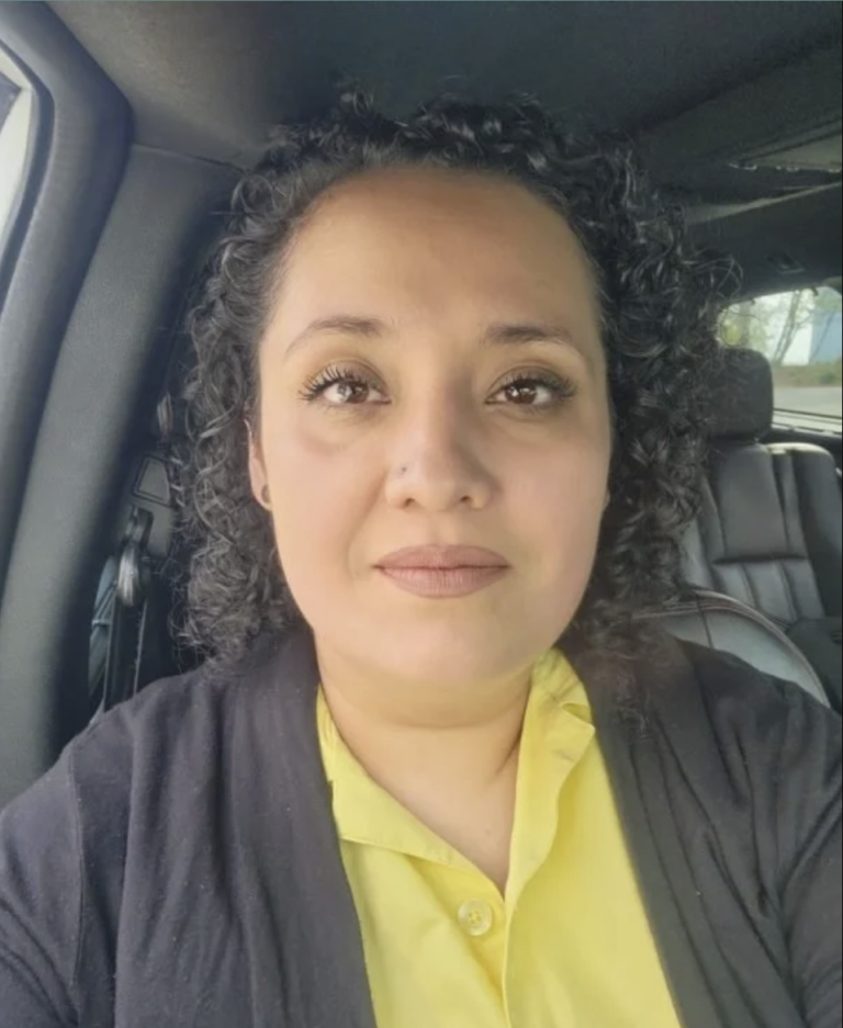 A woman with curly hair is seated in a car, wearing a yellow shirt and dark cardigan.