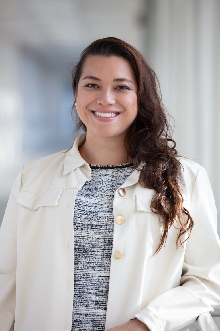 Professional headshot of VPLC employee Blanca Bonilla, who has long, wavy brown hair and is wearing a white jacket with gold buttons and a black and white striped shirt underneath.