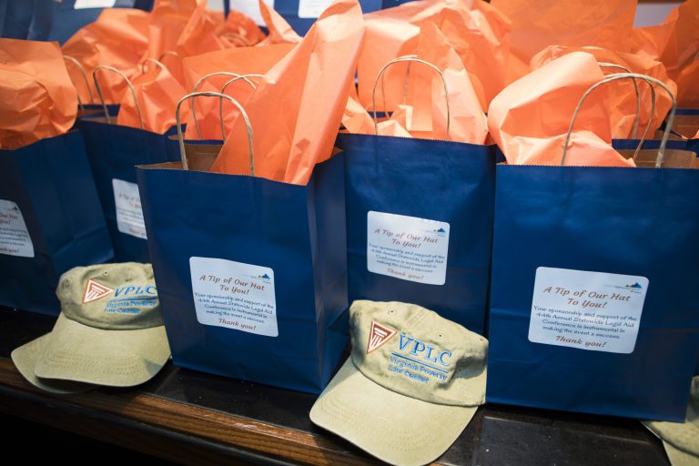 A collection of blue gift bags with orange tissue paper and a green cap featuring the "VPLC" logo, along with a thank-you note for sponsorship and support of VPLC's legal aid conference.