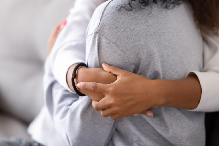 A child and an adult embrace each other warmly, with one wearing a gray sweatshirt and the other a white top. The mood is comforting and supportive.