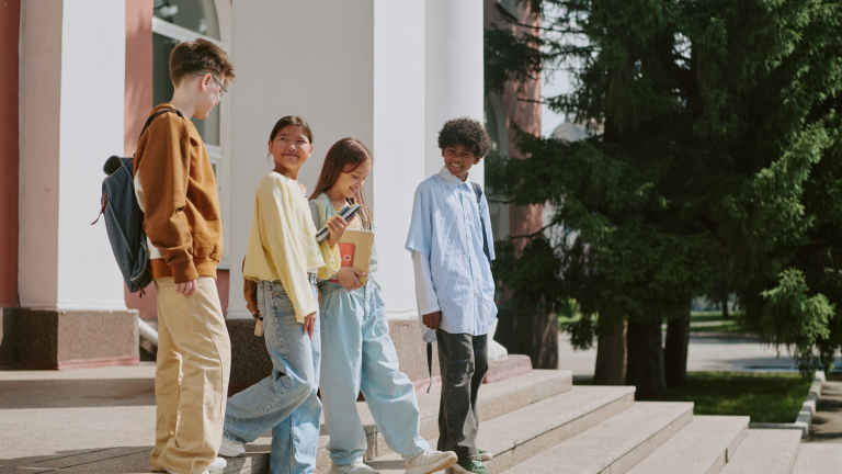 A diverse group of four students chat and smile while standing outside of a building on the steps. They hold books and backpacks, exuding a joyful and friendly vibe.