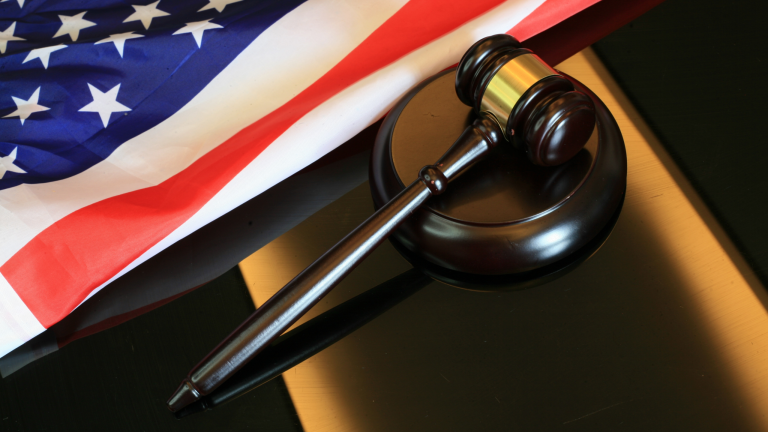A judge's gavel rests on a black surface beside a folded American flag. This image symbolizes justice, authority, and patriotism.