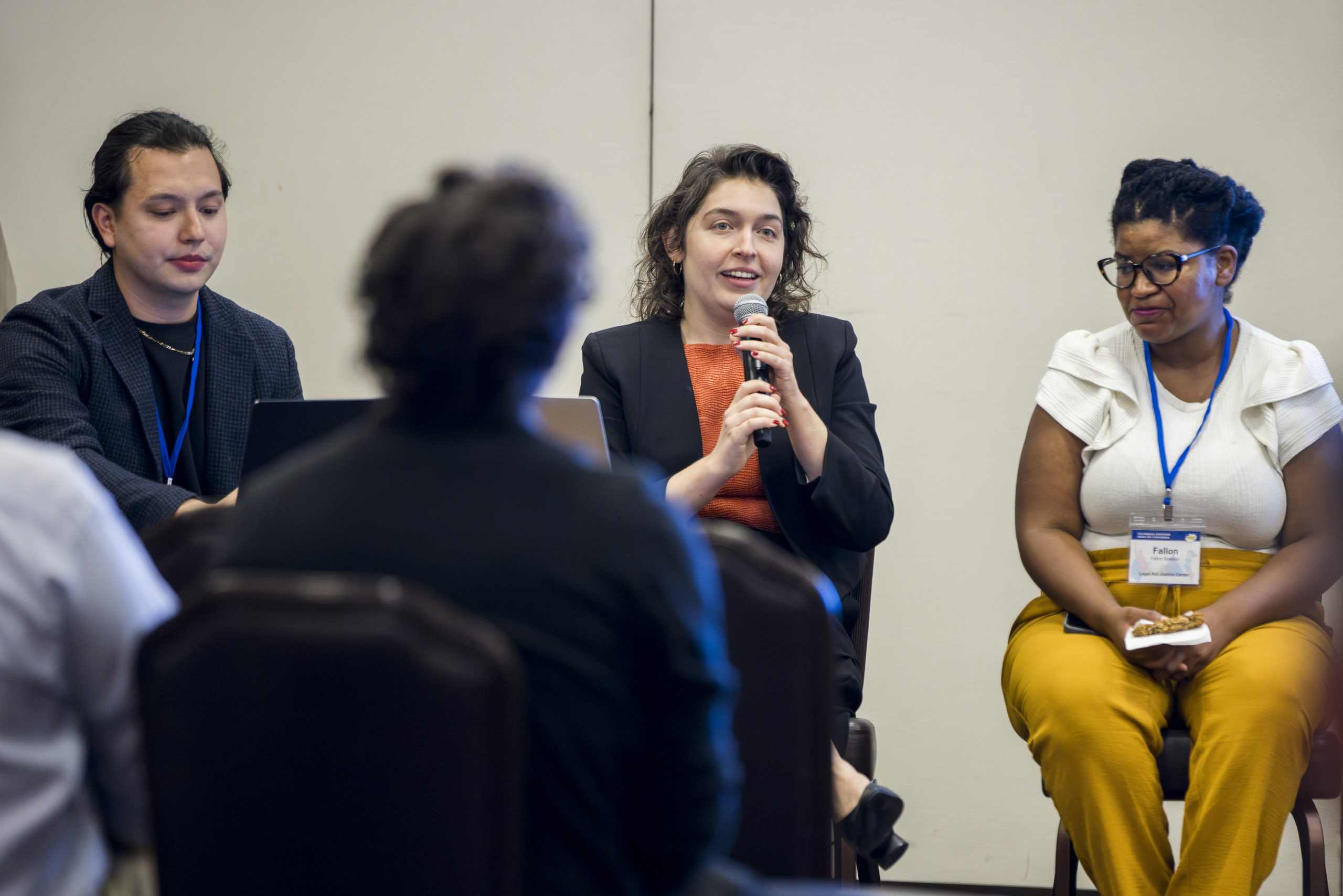 Panel discussion with three speakers; a woman holds a microphone, while attendees listen in a conference room. The mood is engaged.