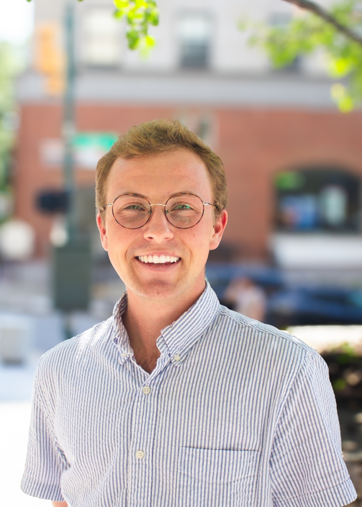 Smiling young man wearing round glasses and a striped button-down shirt, posed outdoors against a softly blurred urban background.