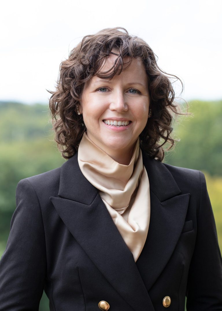 Smiling woman with curly brown hair wears a black double-breasted blazer and beige scarf, standing outdoors against a softly blurred green landscape.