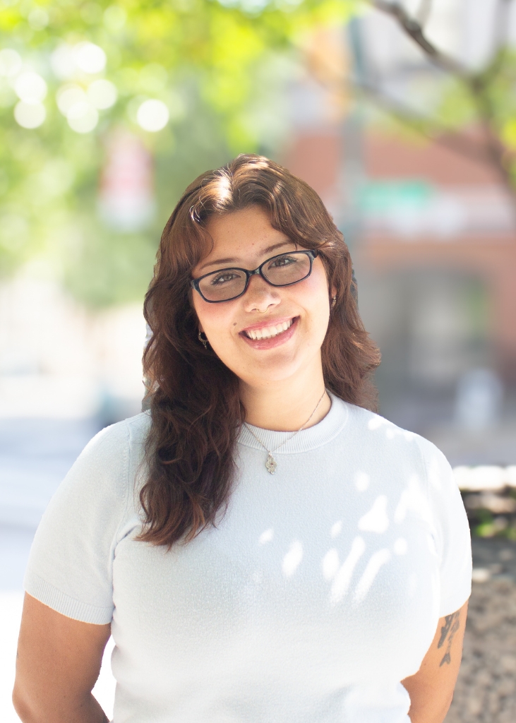 Smiling woman with glasses and long brown hair stands outdoors in dappled sunlight, wearing a white shirt and projecting a warm, friendly mood.