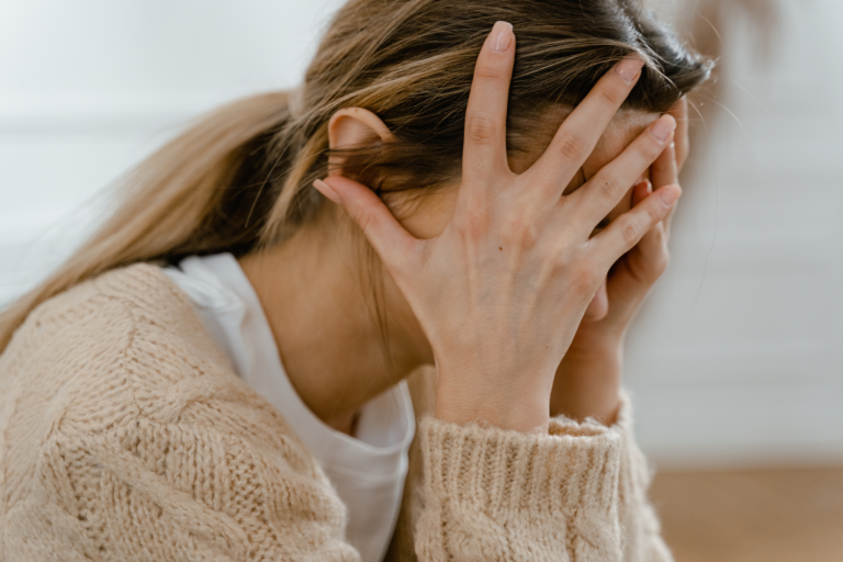 A distressed woman in a beige knit sweater leans forward with both hands covering her face, conveying sadness, stress, or emotional overwhelm.